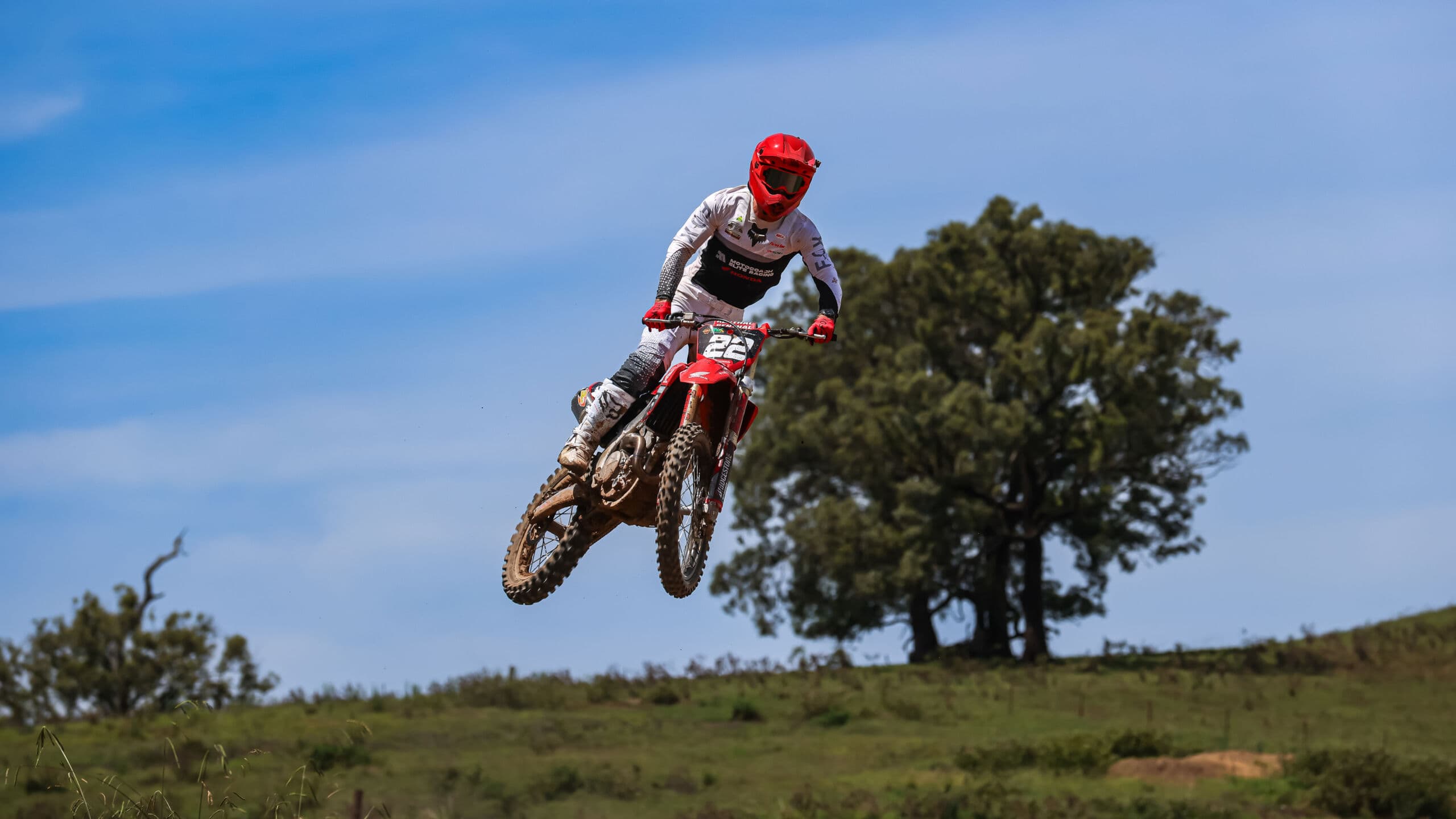 Motocoach Elite Racing rider airborne over a grassy paddock beside a large gum tree
